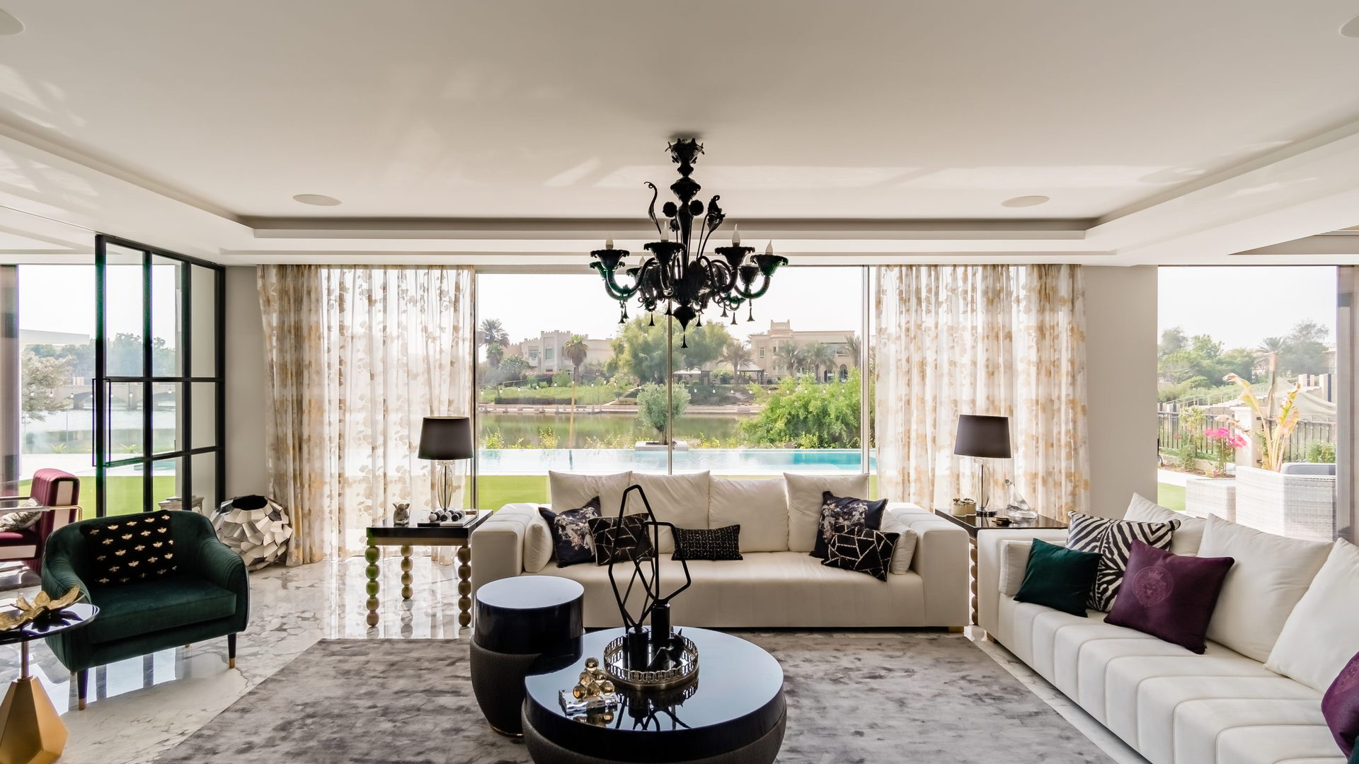 A bespoke residential salon with a bespoke sectional sofa, sculptural centre table and a black crystal chandelier, opening onto a lake view